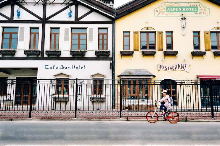 An Asian woman cycling along a street on a  bicycle. The bicycle looks like one that's part of a public bicycle hire scheme. In the background are two buildings, 'Cafe bar hotel' and 'Alpen hotel restaurant', both have similar Alpen architecture. The main difference between the two is the cafe is painted white, whereas the restaurant is painted light yellow