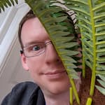 Head-shot of me, a pale white guy, wearing glasses, grinning slightly, and partially hiding behind a plant